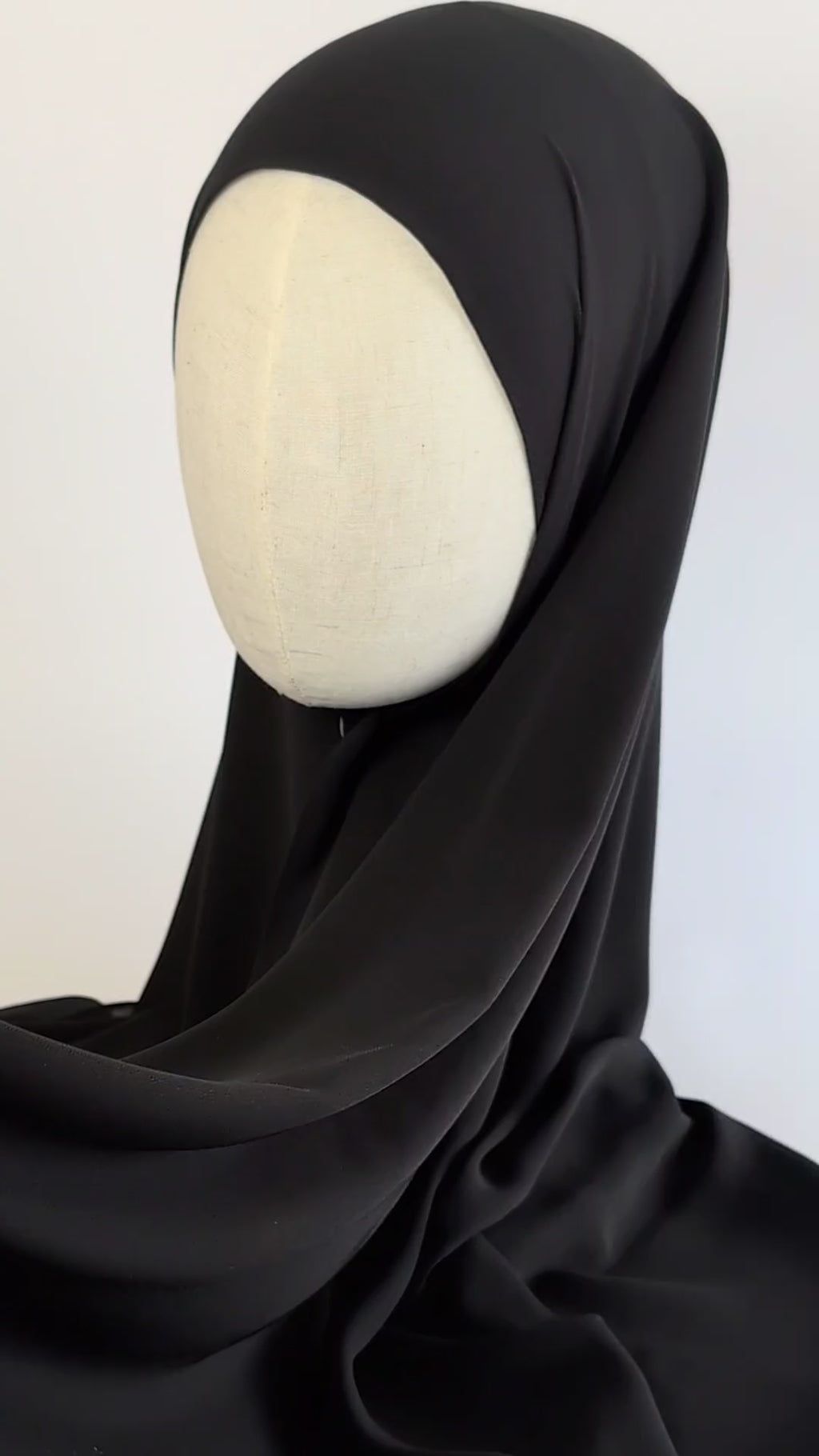 A black chiffon hijab draped over a cream-colored mannequin head in side face shot. the video focuses on how the hijab drapes gracefully and moves.
