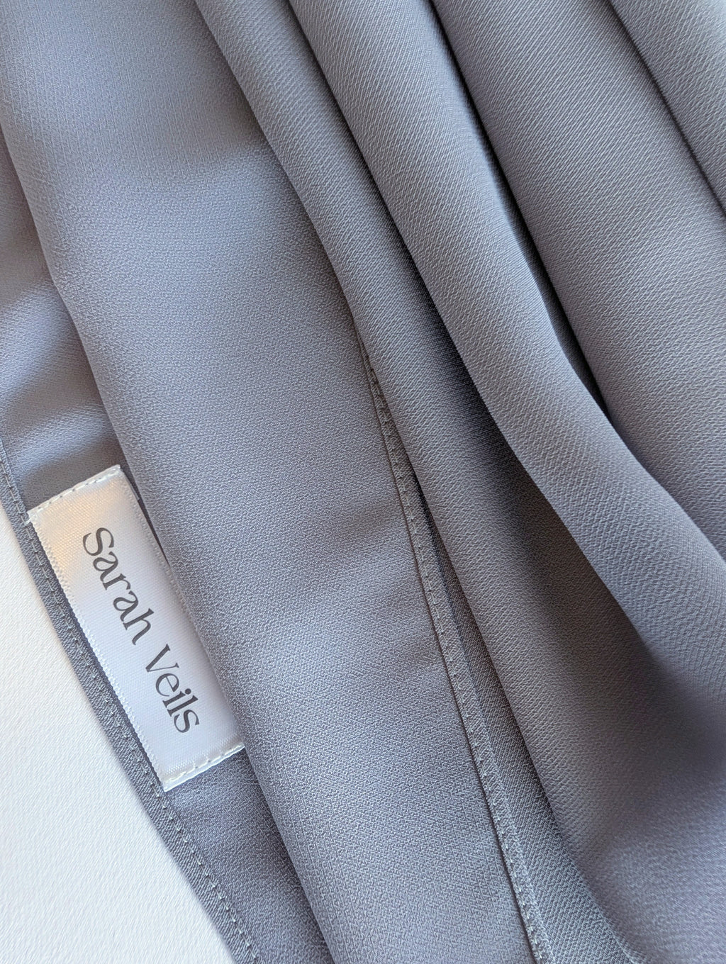 Close-up of a light gray chiffon hijab product with soft folds and a visible white satin label reading Sarah Veils.