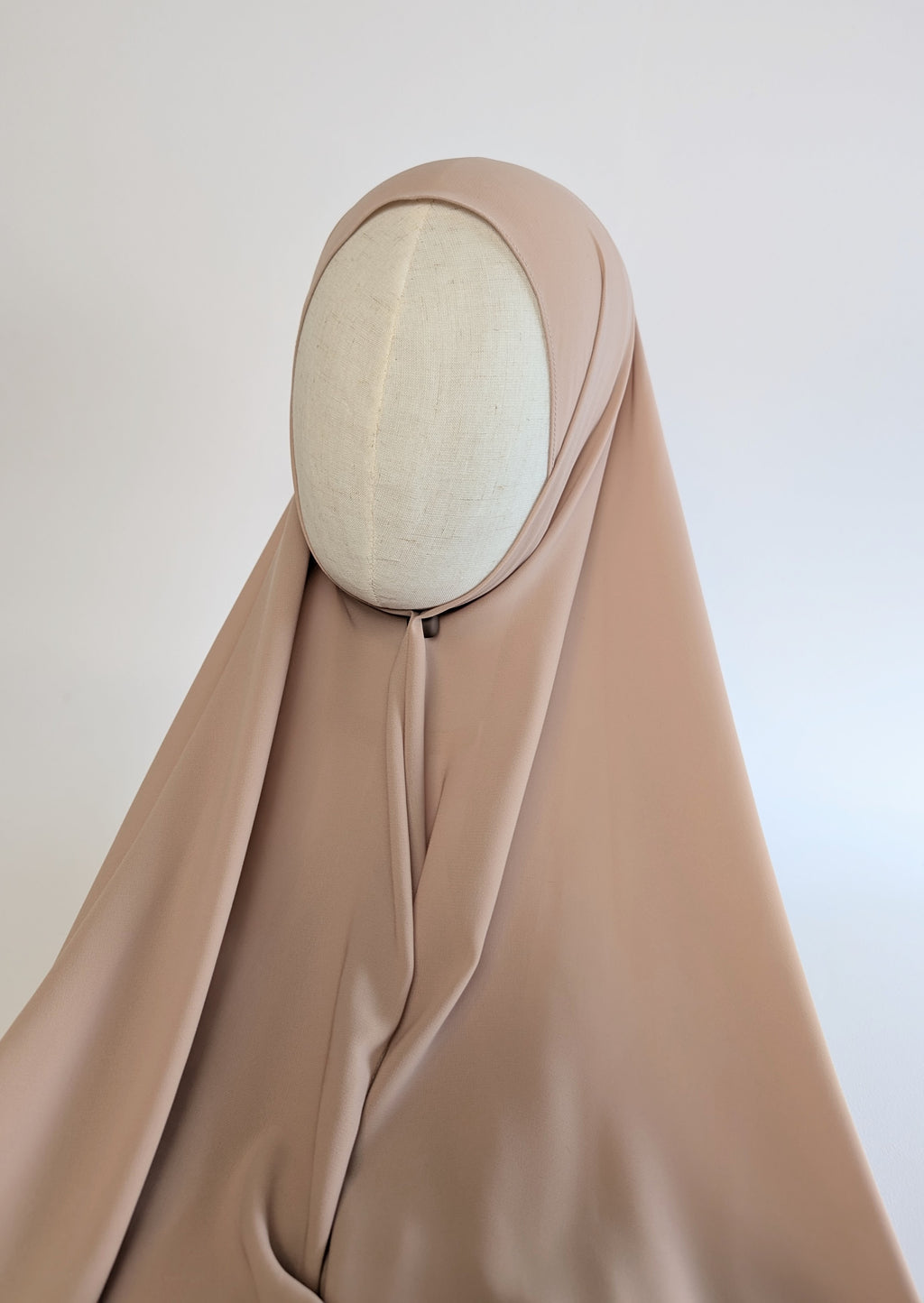 A draped hijab in a soft taupe-beige color, styled on a cream-colored fabric mannequin head to show how it would look on customer in full face front view