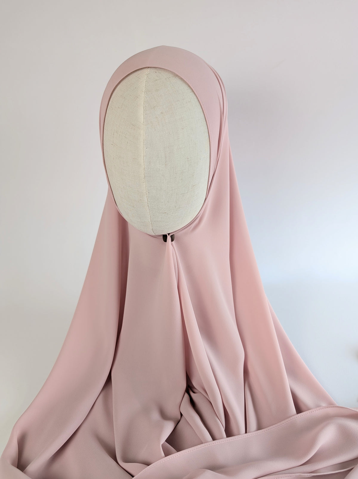 A dusty pink or soft blush pink  hijab draped over a cream-colored fabric mannequin head, set against a plain white background.