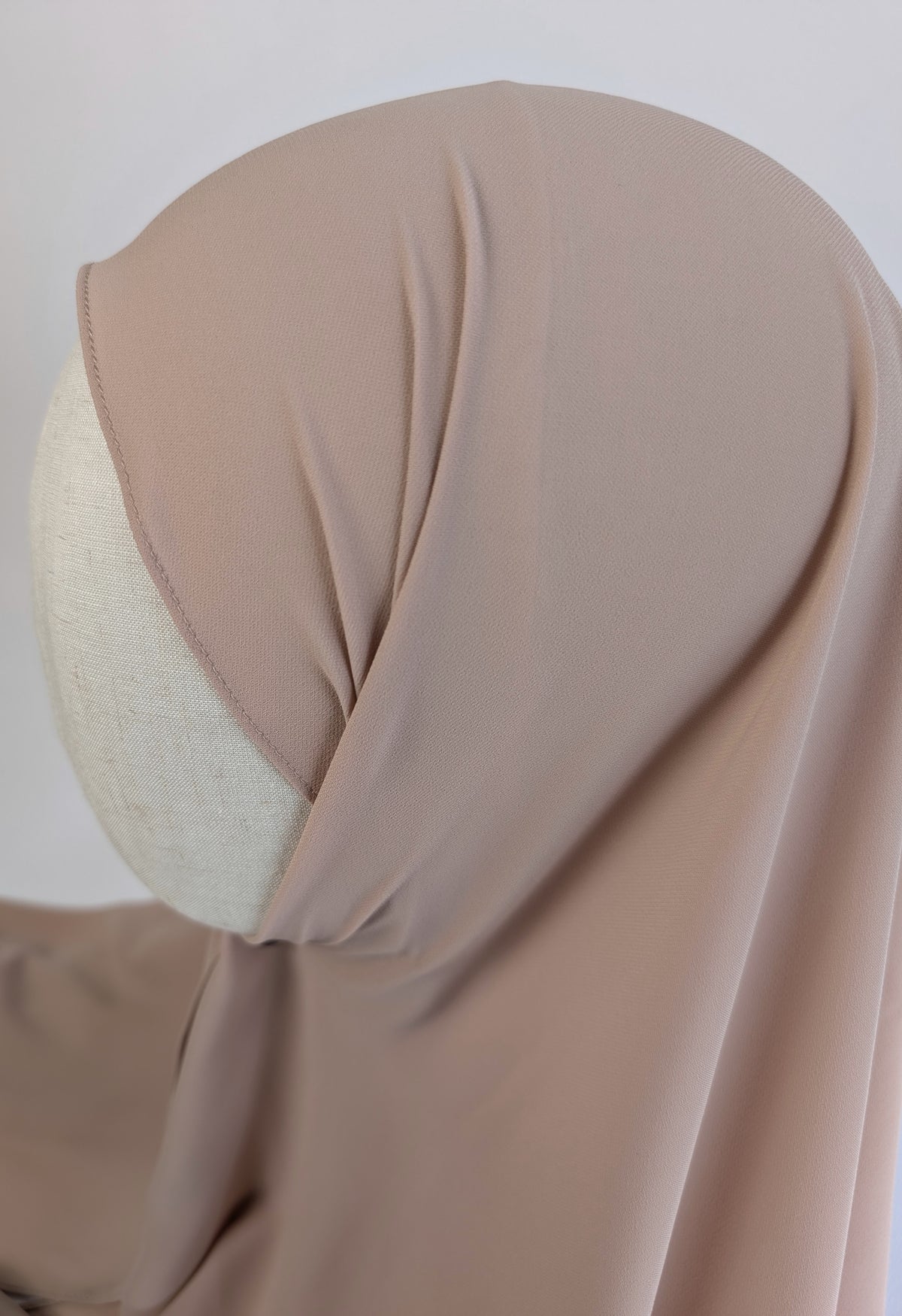 A taupe-beige hijab draped on a cream-colored mannequin head, shown from a side-back angle against a plain light background.