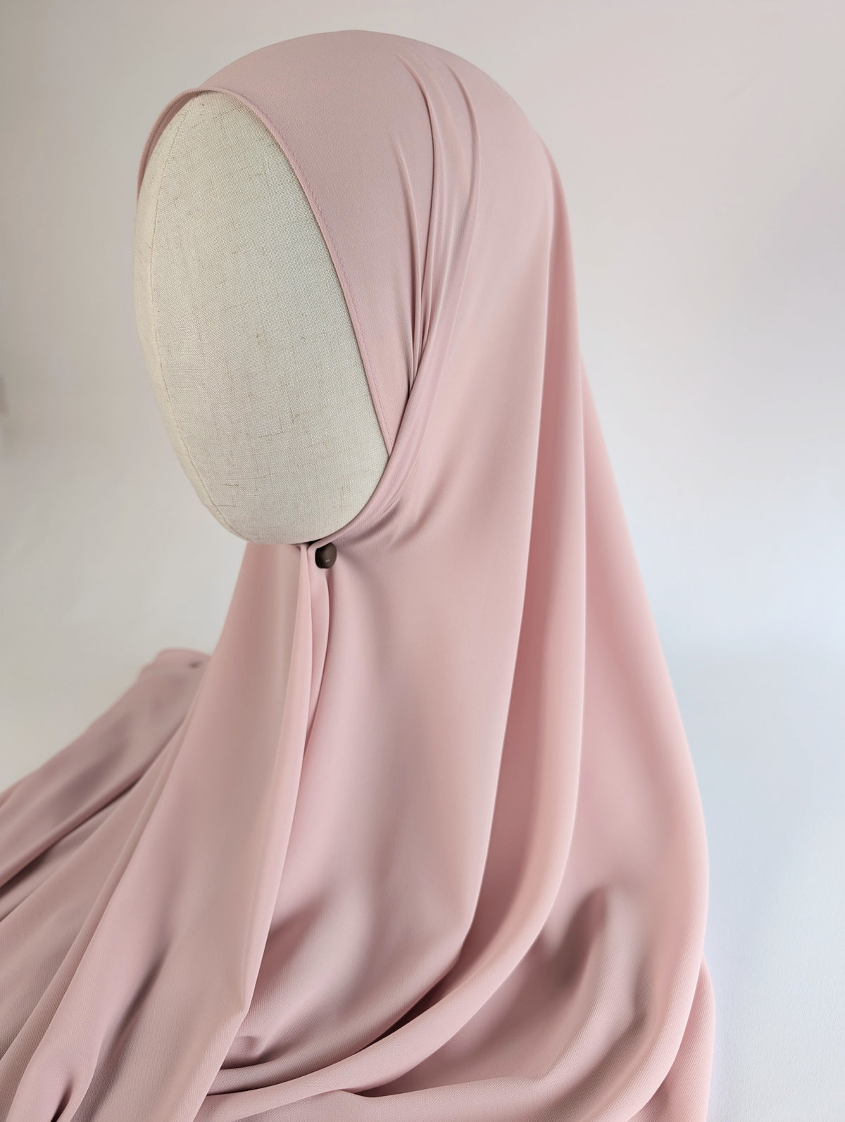 A draped chiffon hijab in a soft dusty pink color, styled on mannequin head model in side face shot against a white background to show how it would fit and look on the customer.