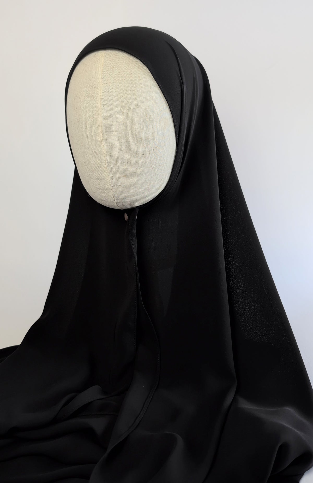 A black draped chiffon hijab/ headscarf or modest garment displayed on a cream mannequin head in full face front shot view against a plain light background